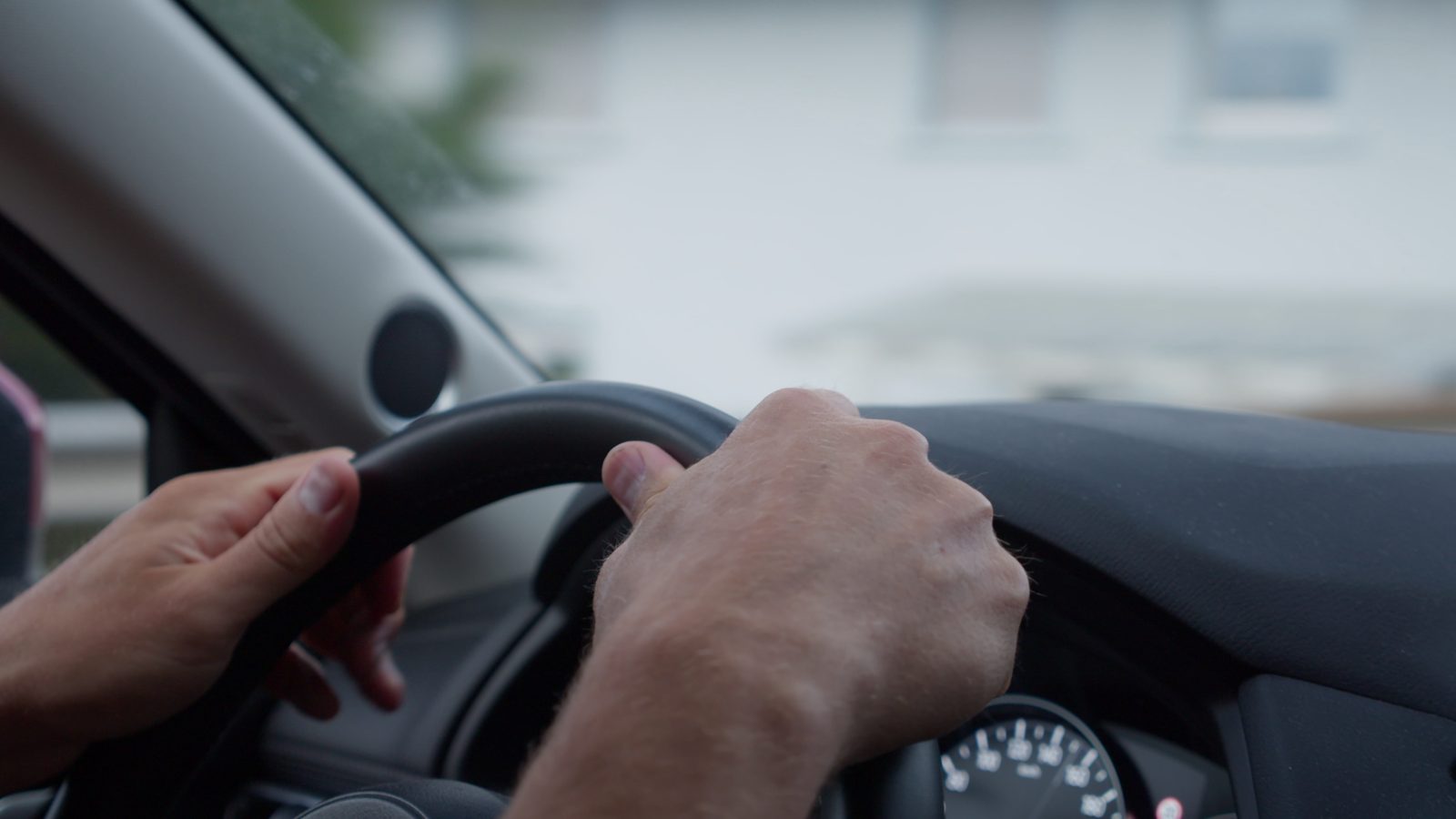 A driver's hands on a car steering wheel, seen from inside the car, with the speedometer below and a blurred street through the windscreen