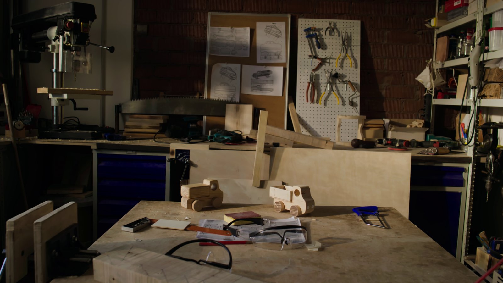 An empty woodworking workshop with a workbench, small wooden toy trucks, safety glasses, a drill press and tools on a pegboard