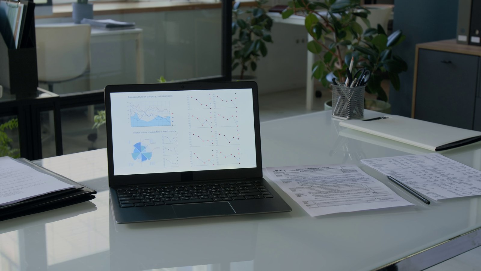 An empty office desk with a laptop showing charts, printed documents and a pen