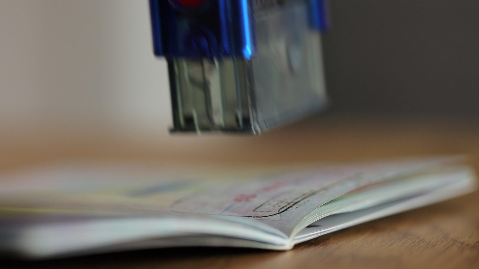 An ink stamp held just above the open pages of a passport lying on a wooden desk